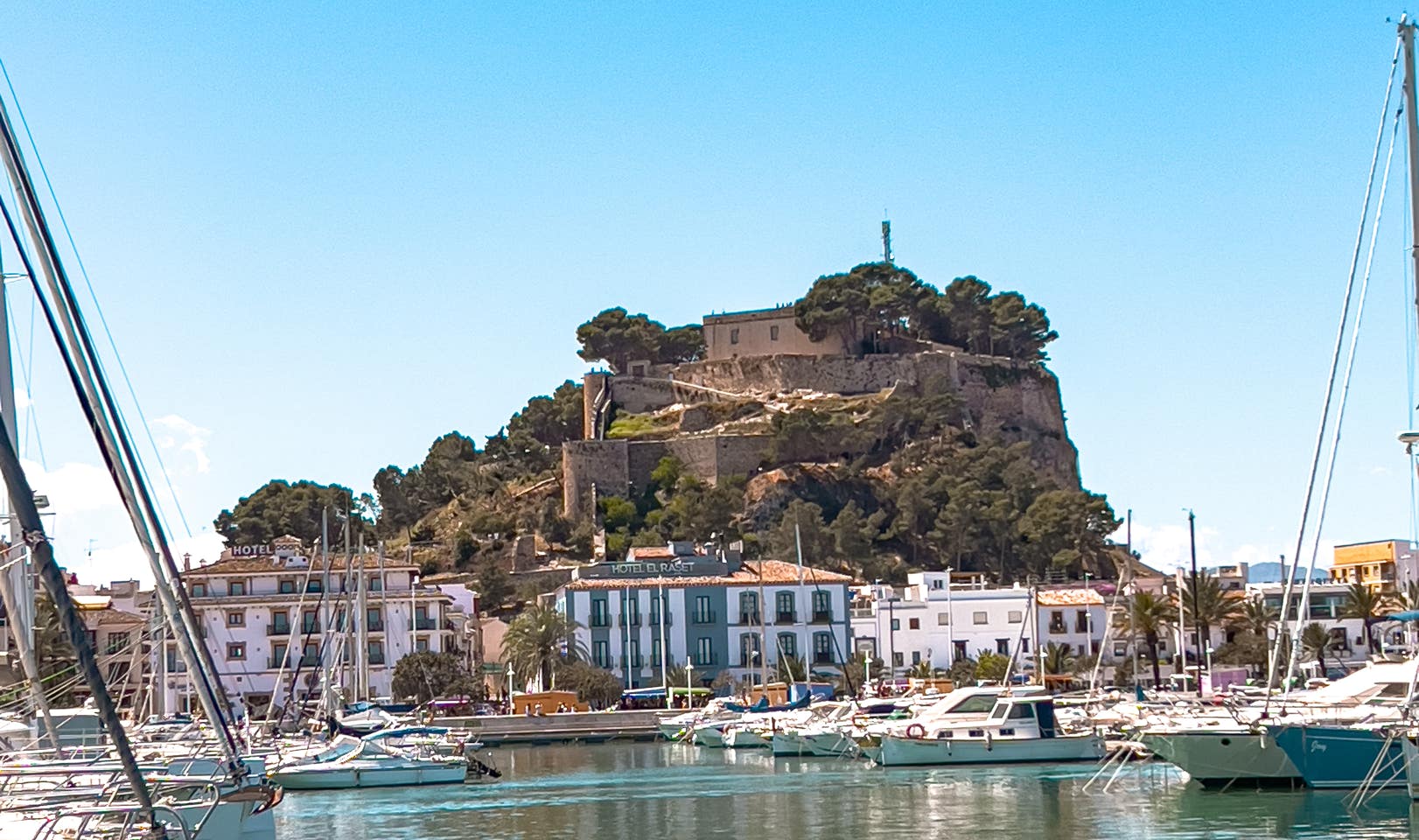 View of the Moorish Dénia Castle rising above the marina, surrounded by whitewashed buildings and sailboats on the sunny Mediterranean coast