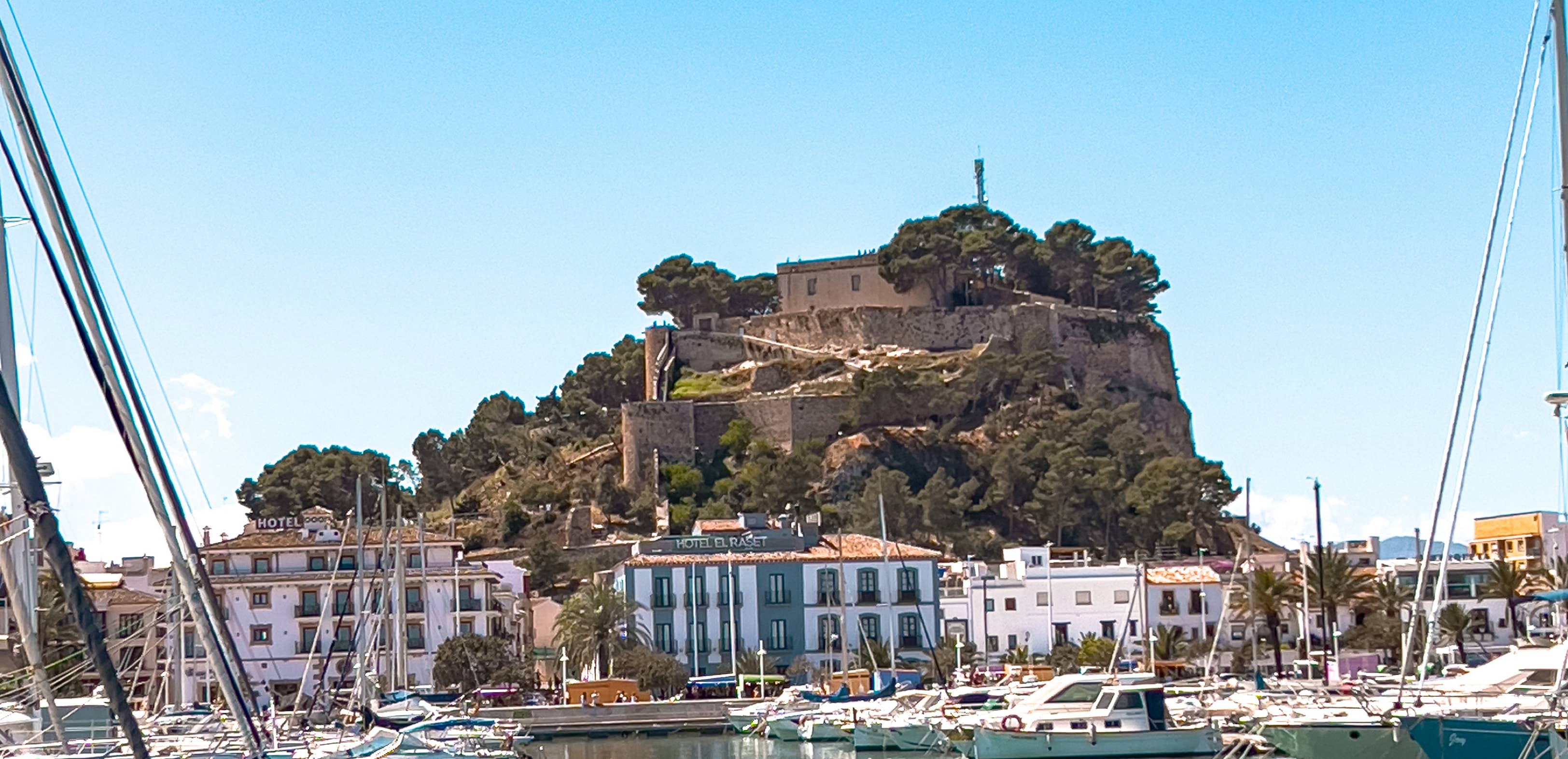 View of the Moorish Dénia Castle rising above the marina, surrounded by whitewashed buildings and sailboats on the sunny Mediterranean coast